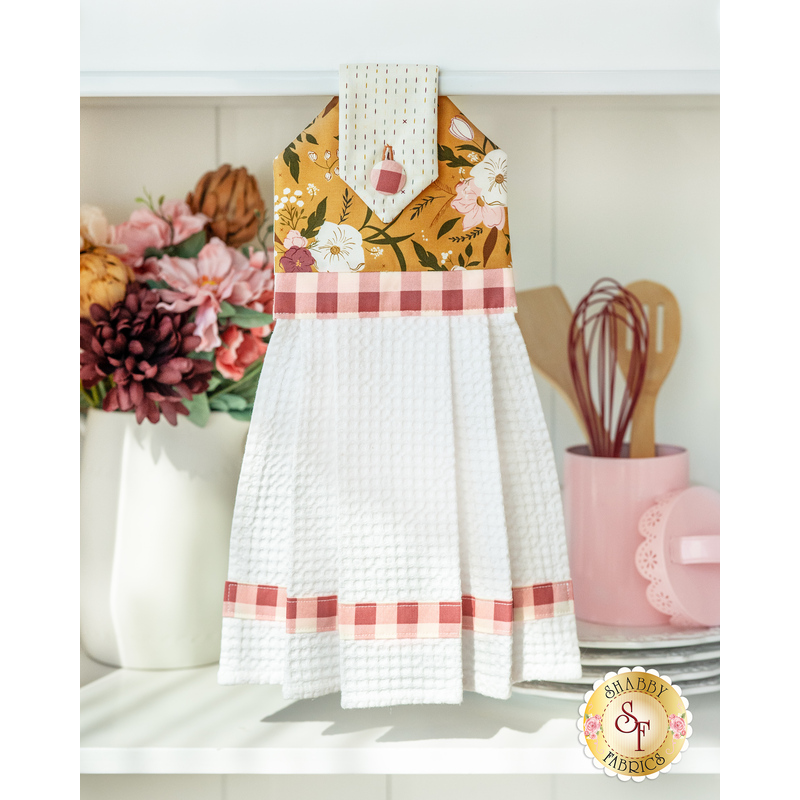 Photo of a white kitchen towel with floral and gingham fabrics with vases and crocks in the background holding flowers and kitchen utensils