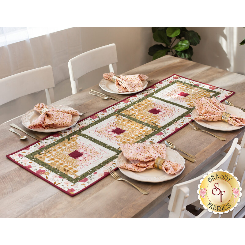 Photo of a table runner made with log cabin blocks and cream, yellow, green, and red floral fabrics laying in the center of a table with light wood and white chairs and four place settings at each seat with matching napkins