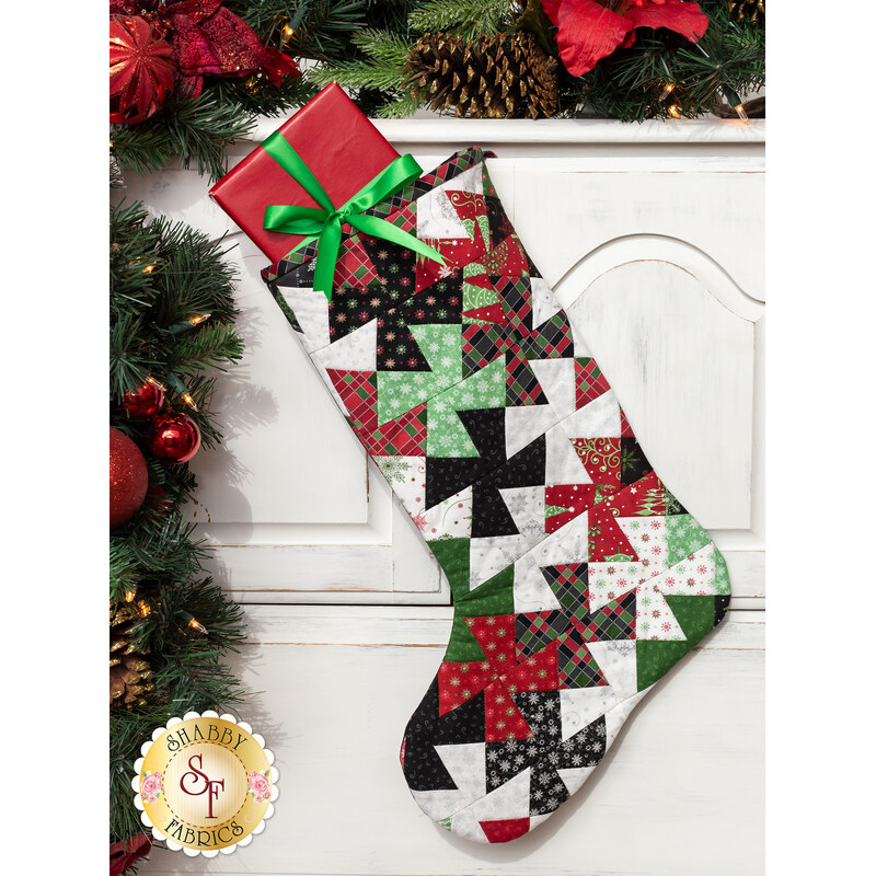 Photo of a Christmas stocking made with red, green, black, and white Christmas fabrics hanging from a white mantle with garland all around and a red wrapped gift inside the stocking.