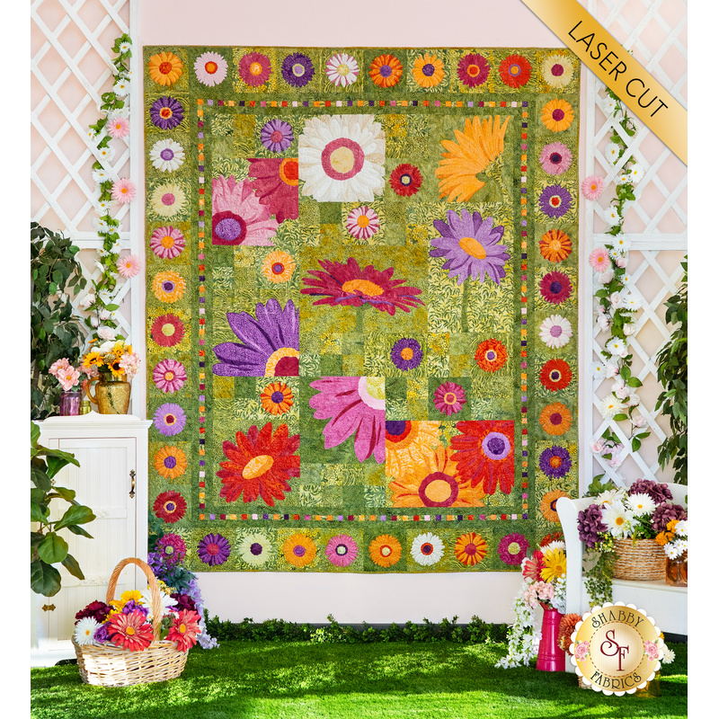 Photo of a green, pink, purple, and yellow floral quilt hanging on a white paneled wall with white lattices on either side with baskets of colorful flowers and climbing plants