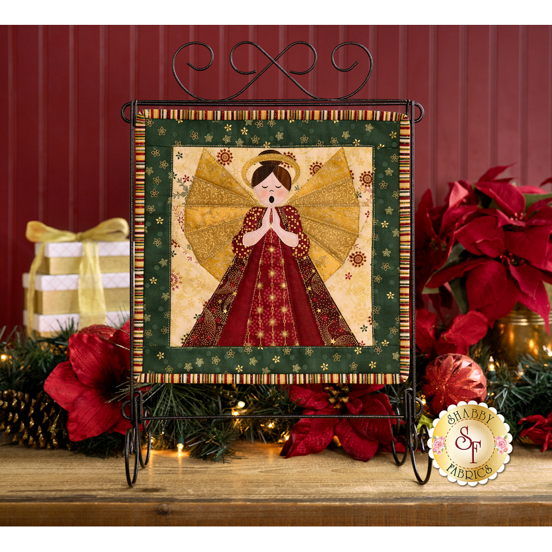 Photo of the finished FPP project for December featuring a praying angel and hanging on a tabletop craft holder with Christmas decor in the background in front of a maroon paneled wall