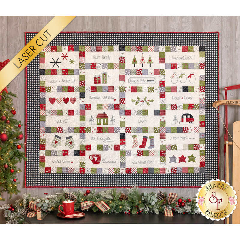 Photo of the Hometown Christmas quilt hanging flat on a gray wooden wall with Christmas decor all around like a wooden sled, decorated Christmas tree, and a mug of hot cocoa.