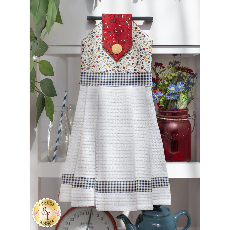 The completed hanging towel in white and red with a yellow fabric button and stripes of navy blue gingham. It is hung on a drawer and staged with coordinating decor.