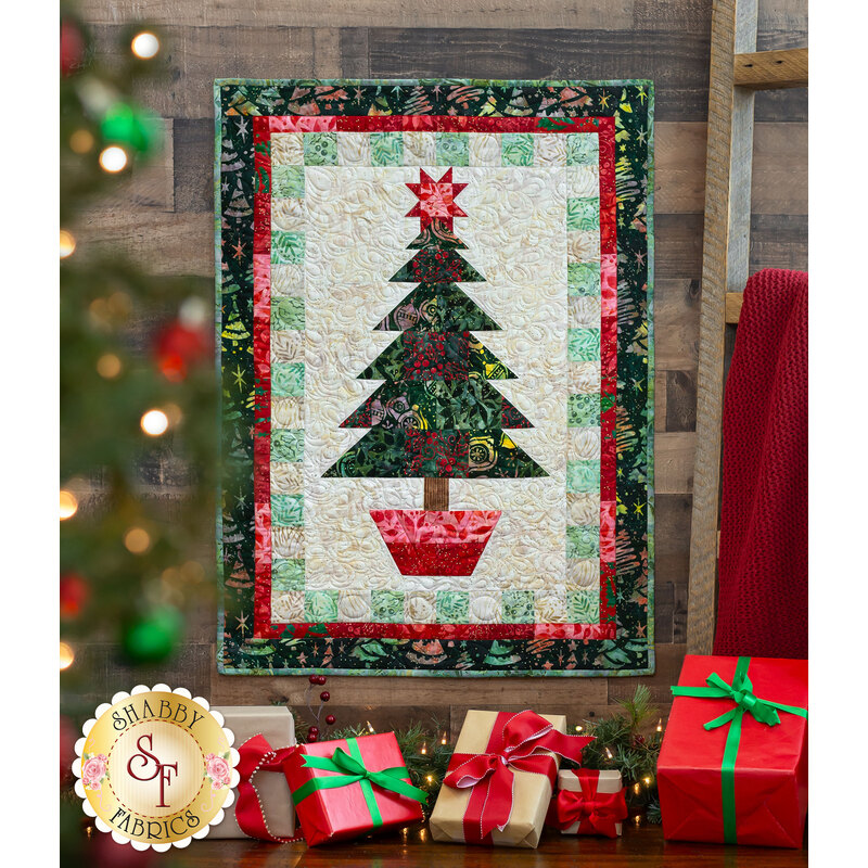 Photo of a Christmas quilt featuring a large potted pine with a star on top, hanging on a brown paneled wall with a ladder and red blanket to one side, a decorated Christmas tree in the foreground, and wrapped gifts all along the floor
