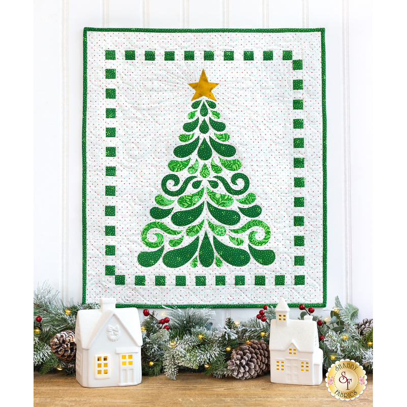 Photo of a Christmas tree wall hanging on a white paneled wall with winter garland and tiny decorative houses on a countertop below