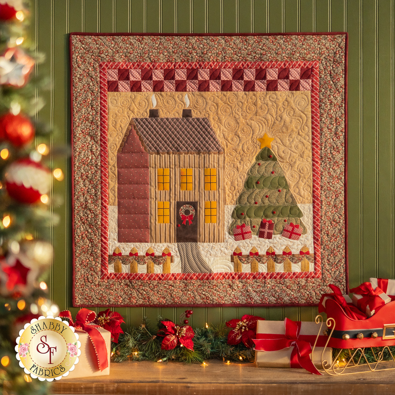 A cream paneled wall with a small wall hanging depicting a house in a snowy field with a Christmas tree in the yard decorated with wrapped gifts. Christmas decor is all around the wall hanging with a decorated tree in the foreground.