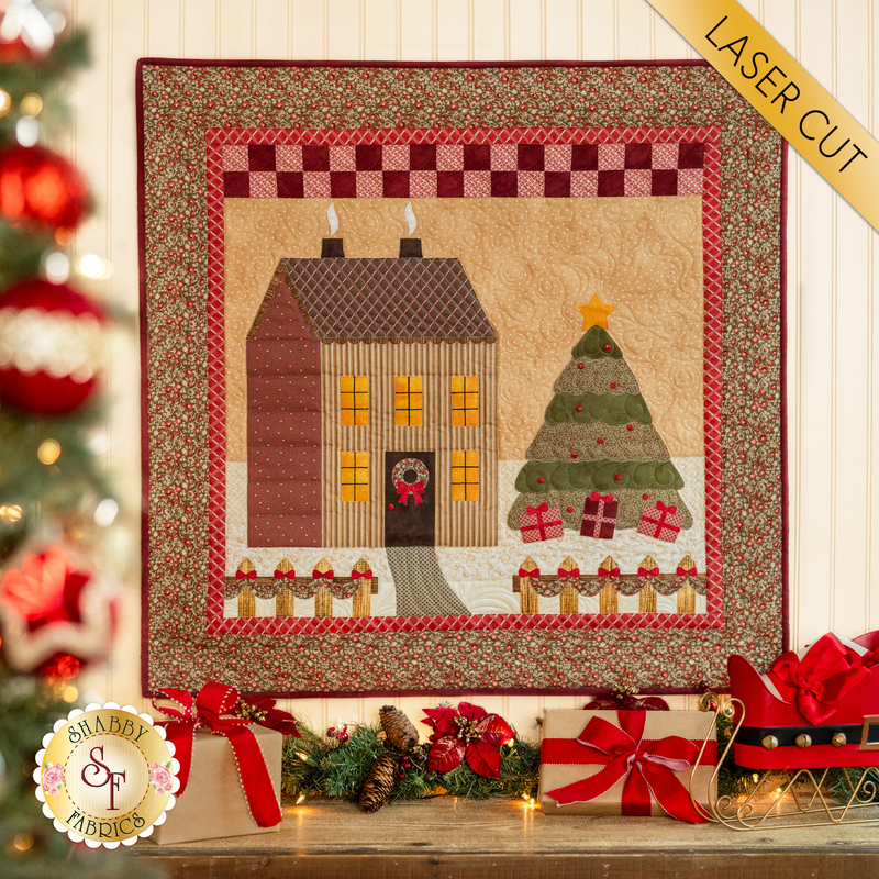 A cream paneled wall with a small wall hanging depicting a house in a snowy field with a Christmas tree in the yard decorated with wrapped gifts. Christmas decor is all around the wall hanging with a decorated tree in the foreground.