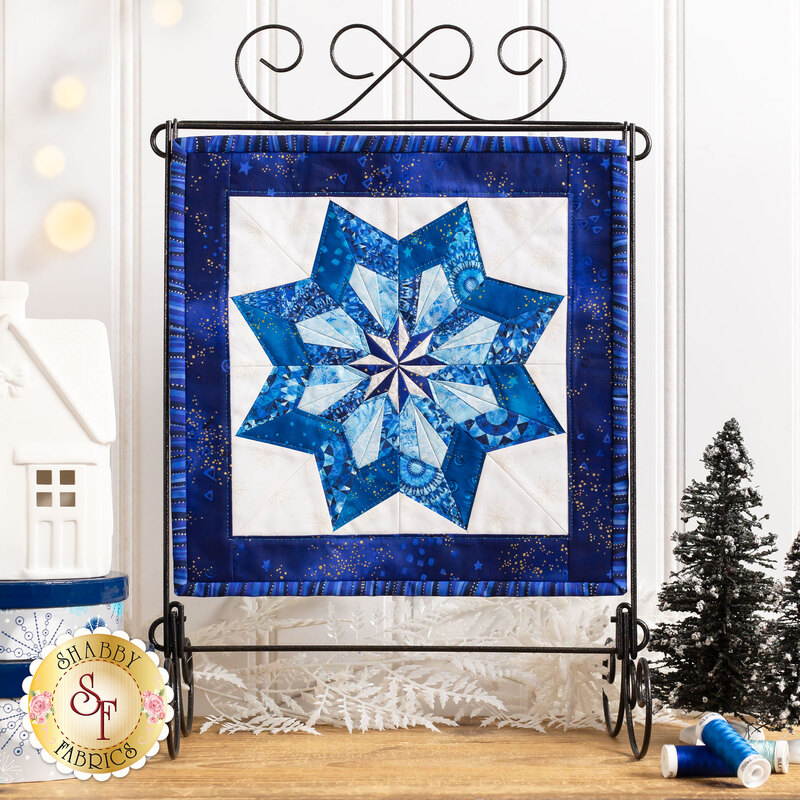 Photo of the completed quilt block featuring a pinwheel shaped snowflake made with different shades of blue fabric with a white background and dark blue border hanging on a tabletop craft holder atop a wooden counter with winter decor.