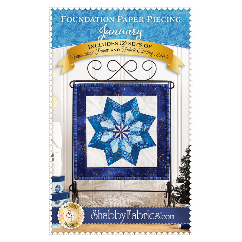 Front cover of the pattern booklet featuring a photo of the completed quilt block featuring a pinwheel shaped snowflake made with different shades of blue fabric with a white background and dark blue border hanging on a tabletop craft holder atop a wooden counter with winter decor and including designer and pattern name info.