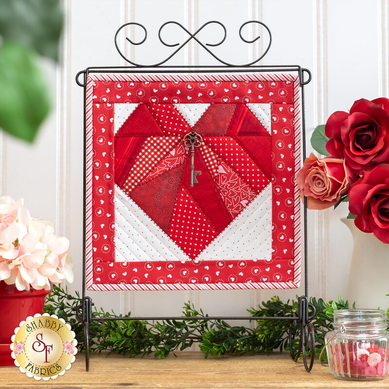 Photo of the completed February project featuring a red heart and key charm on a white background with a red border hanging on a craft holder in front of a white paneled wall with floral decor on either side.