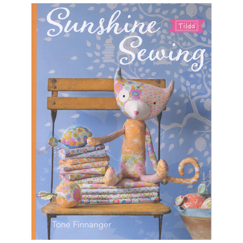 Front cover of the pattern book showing a little cat doll made of bright fabrics, staged on a vintage chair with an array of bright fabrics and a basket of fabric fruits