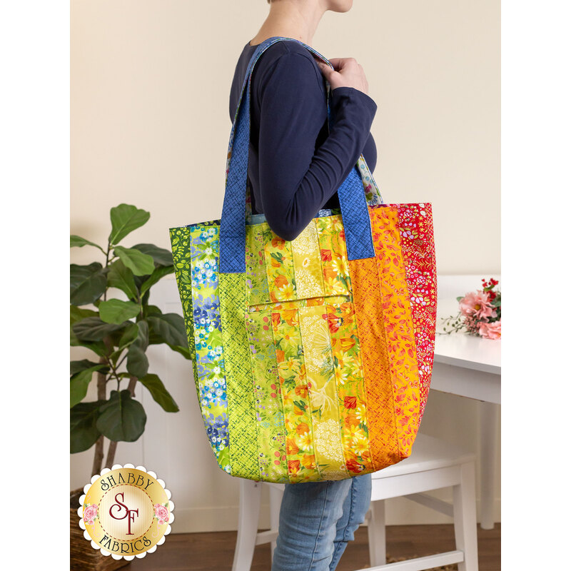 Photo of a large jelly roll tote made with bright floral fabrics slung over a woman's shoulder with a white chair, bouquet, and table in the background