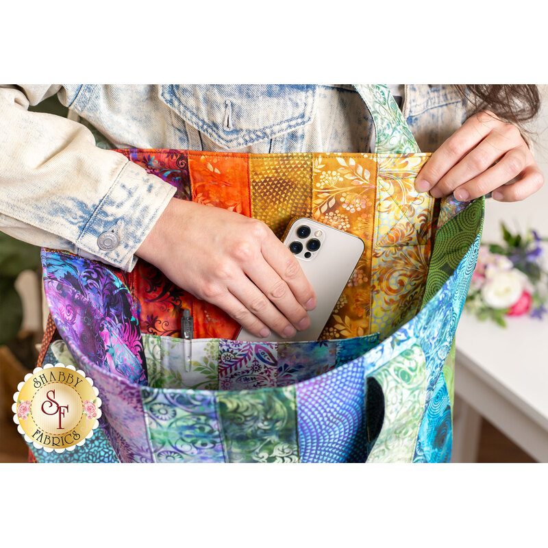Photo of a large jelly roll tote made with purple and blue fabrics over the shoulder of a woman reaching into the inside pocket to pull out a white cell phone with a white table, bouquet, and house plant in the background.