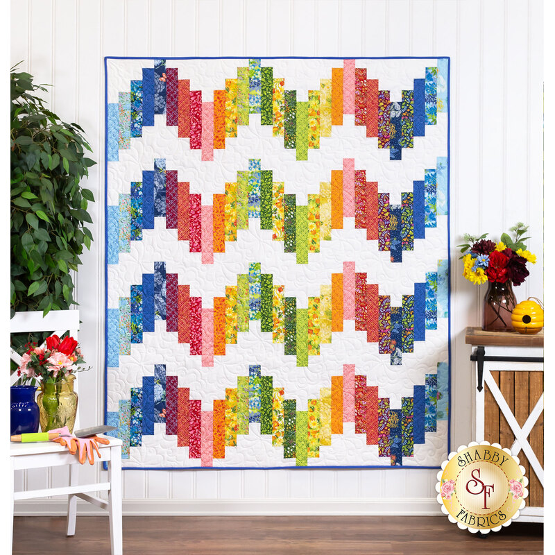 The completed Ridiculously Easy Jelly Roll Quilt in Wild Blossoms, staged on a white paneled wall beside a house plant, furniture, and flowers in coordinating colors.