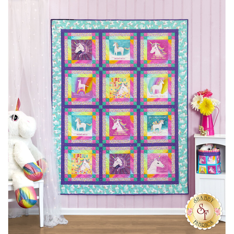 The completed Unicorn Love quilt, staged on a pink paneled wall beside unicorn themed stuffies and color coordinated furniture.