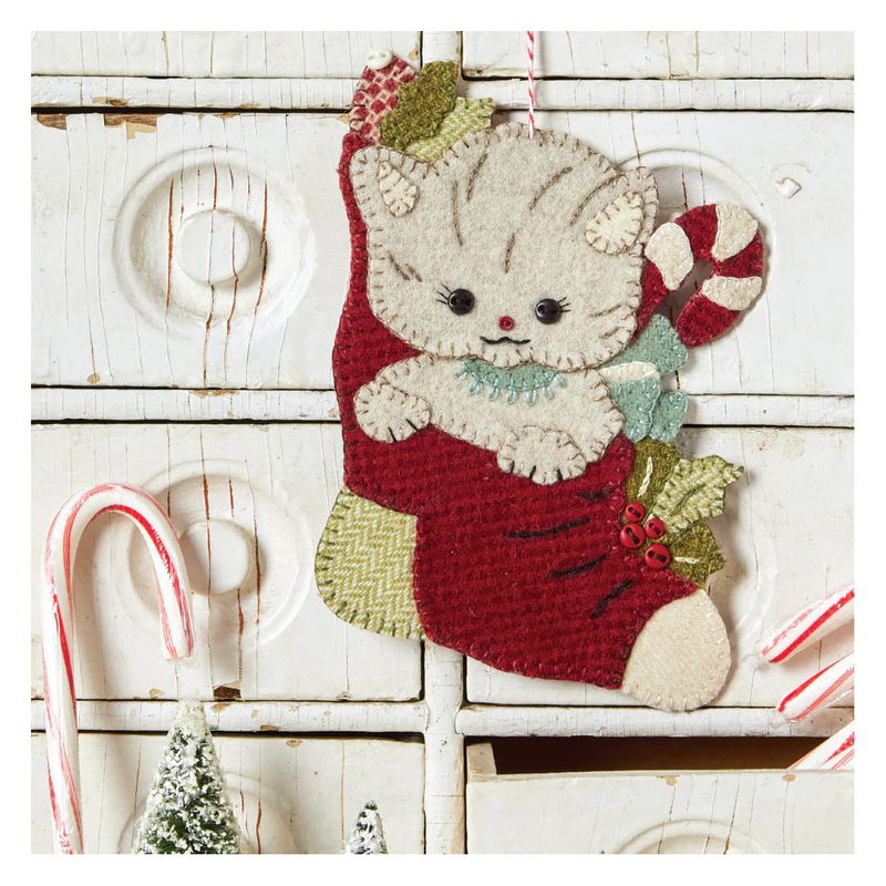 Front cover of pattern, showing the completed kitty ornament, staged over rustic wooden drawers and surrounded with candy canes and tiny model trees.