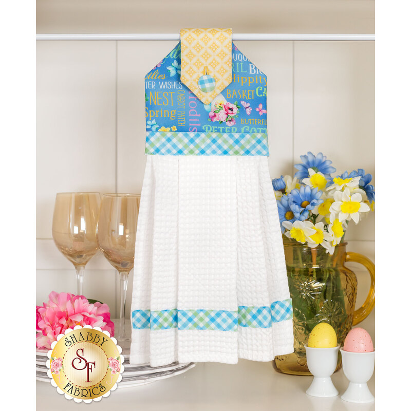 The completed hanging towel in blue, staged on a white counter among coordinated kitchen wares like glasses, plates, and egg cups, as well as a bouquet of yellow and blue flowers.