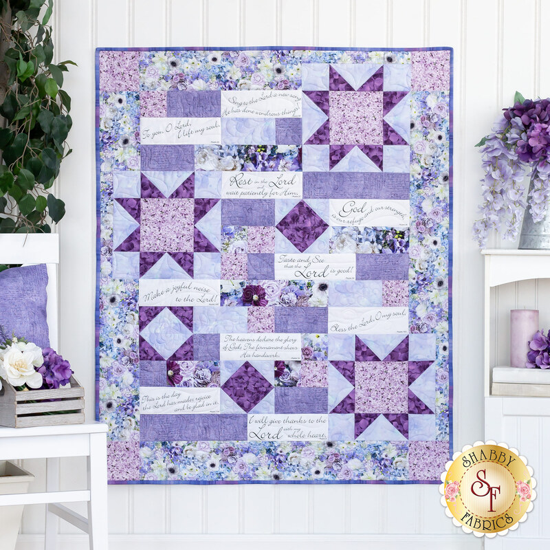 Photo of a purple, blue, and white quilt made with a psalms panel hanging on a white paneled wall with white furniture and purple floral decor on either side.