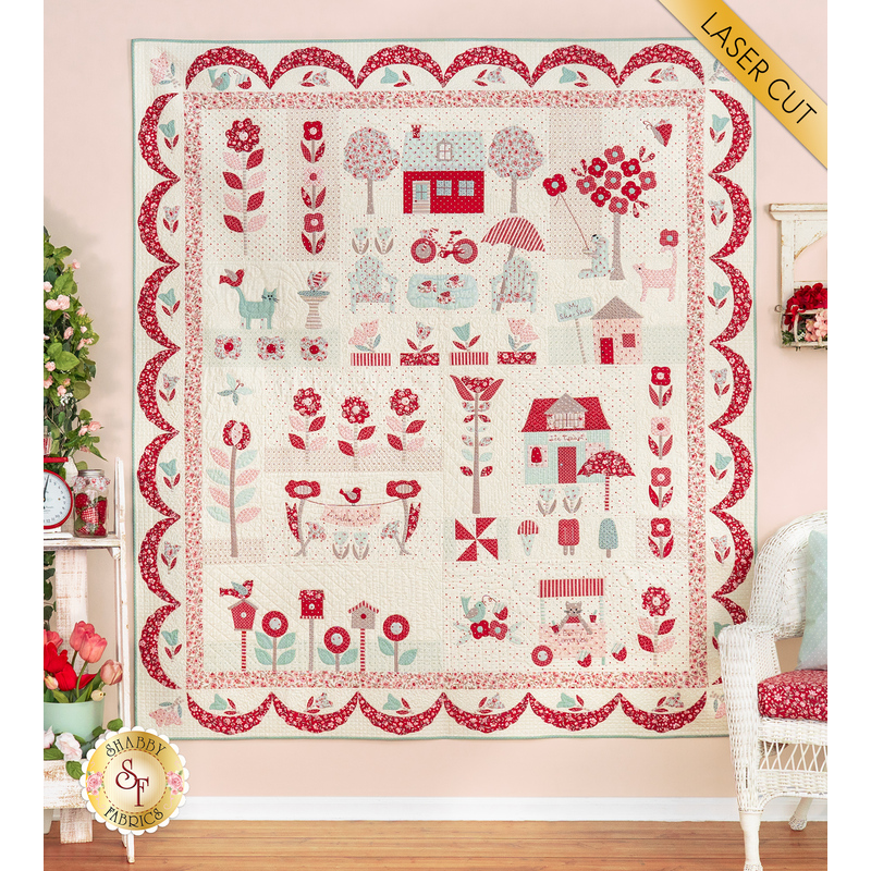The completed quilt in cream, red, and pale teal blue, hung on a pink paneled wall and staged with coordinating furniture. A golden banner reads 