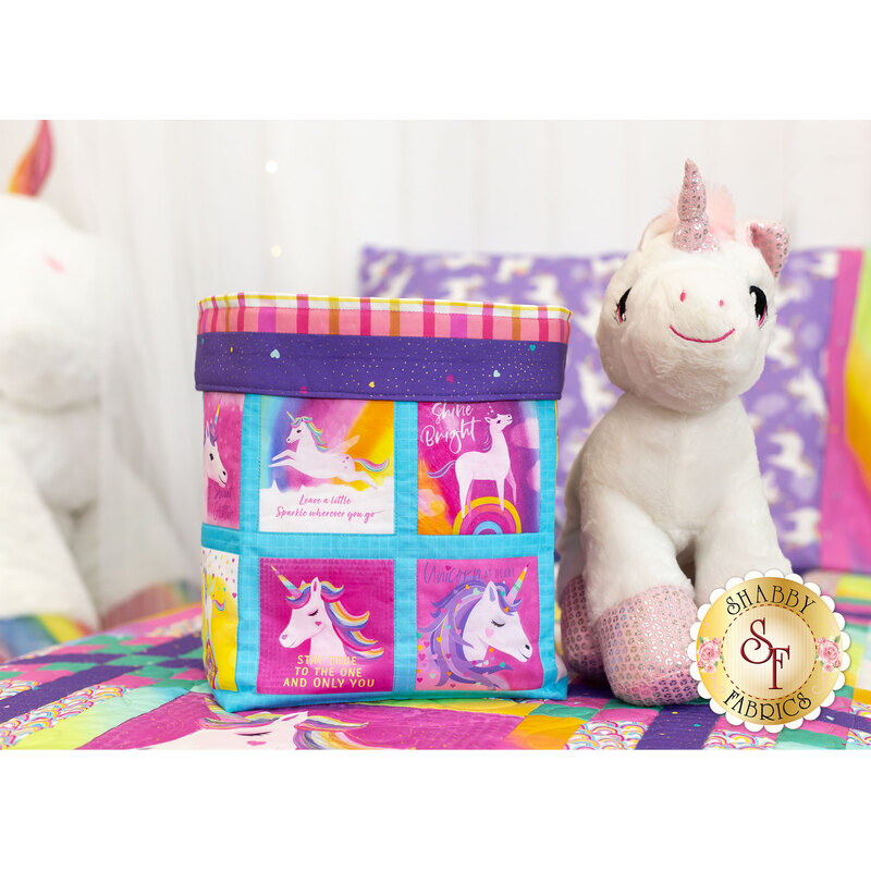 The completed multicolor fabric basket, staged on a bed with the Unicorn Love quilt, the purple magic pillowcase, and two unicorn stuffed animals.