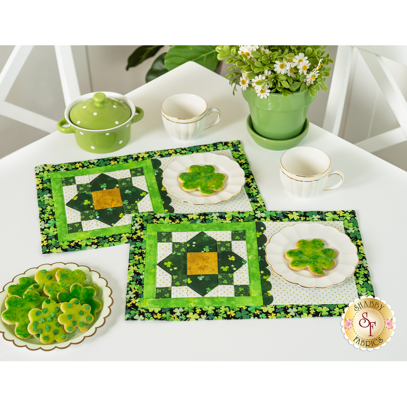 The two completed placemat sets in green and gold, staged on a white table with plates of shamrock cookies, coordinating green house wares, and flowers.