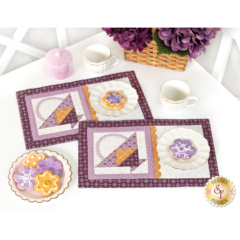 The two completed Tea & Cookies placemats for May, colored in rich purples, gold, and cream, staged on a white table with white teacups and coordinating cookies. A basket of purple flowers peeks into frame from above.