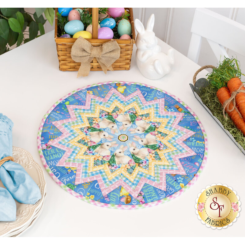 The completed Cottontail Farms Table Topper, colored in blue, yellow, and pink, with a nest of bunnies at the center, staged on a white table and surrounded with coordinated wares like a basket of Easter eggs and a bundle of orange carrots.
