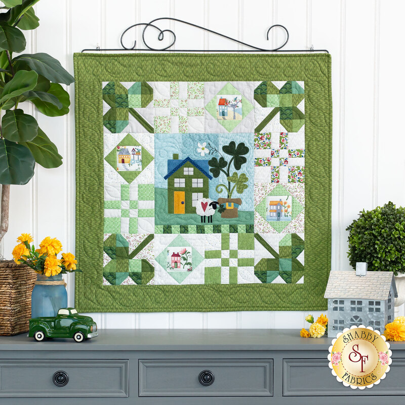 The completed Shamrock Ridge wallhanging, hung on a spiral craft hanger on a white paneled wall, staged with houseplants and coordinating decor.