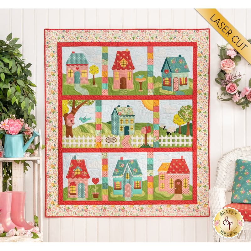 The completed Welcome Home In Spring quilt in bright, cheerful colors, hung on a paneled wall beside pink rainboots, a watering can planter with pink flowers, and a wreath of pink roses. A yellow banner in the upper right hand corner reads 