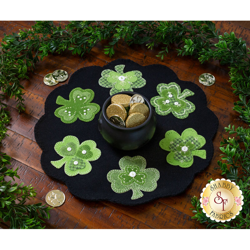 The completed wool mat, staged on a rustic cherry wood table, surrounded by a green garland and tossed golden coins. A small black cauldron filled with sparkling gold medallions sits in the center. 