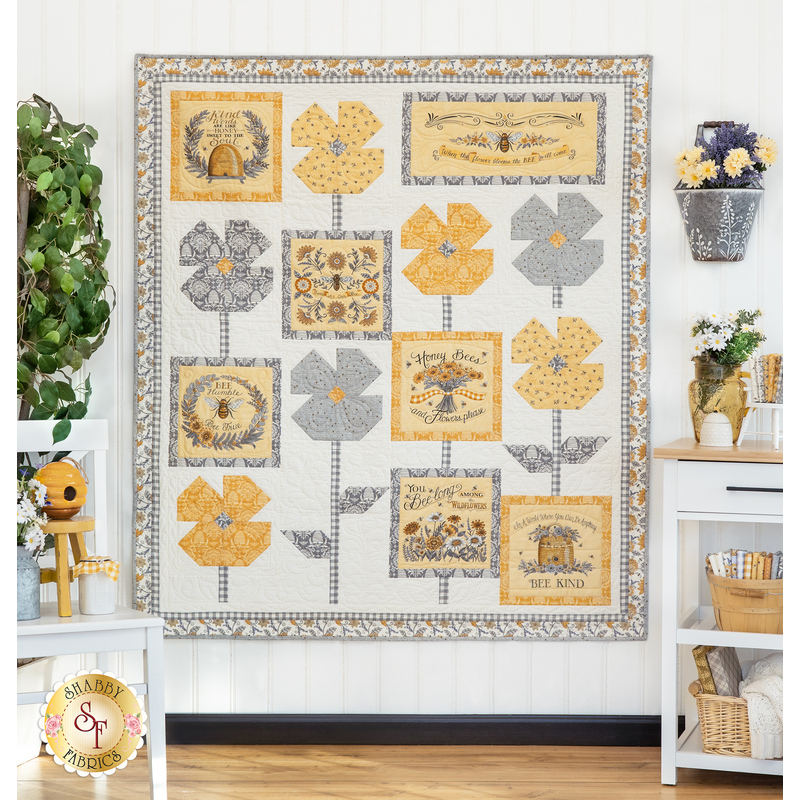 The completed Bees In The Garden quilt, colored in yellow, gray, and cream, hung on a white paneled wall beside coordinating furniture and decor.