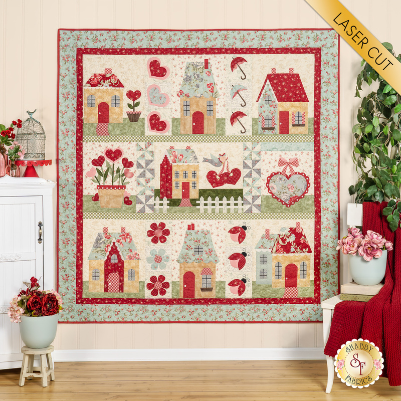 The completed English Cottages quilt, colored in warm reds, soft teal, and classic cream, with mossy green and baby pink accents. It is staged on a cream wall and bordered by coordinating furniture and housewares. A yellow banner at the top right reads 