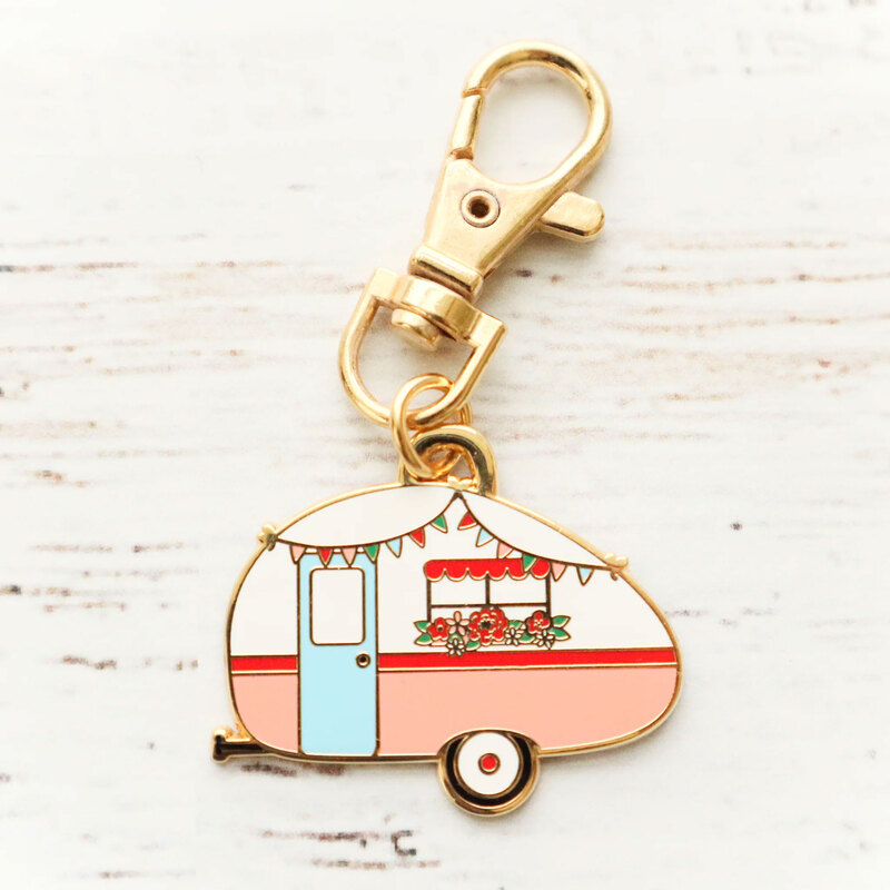 The retro camper enamel charm out of its packaging, staged on a textured white wood background.