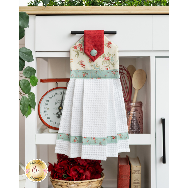 The completed hanging towel in cream, red, and blue, staged on a kitchen cart with cooking utensils, a scale, flowers, and books.