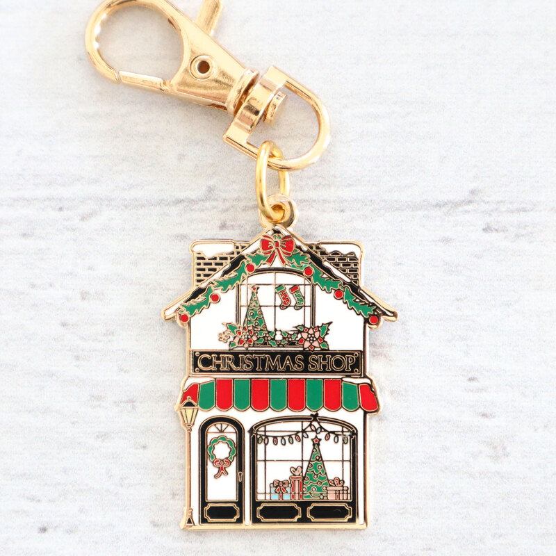 The Christmas Shop enamel charm out of its packaging, staged on a textured white wood background.
