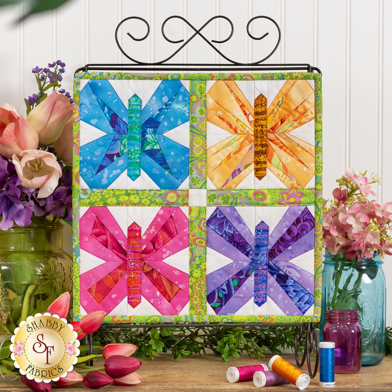 The completed April wallhanging in bright and cheerful blue, yellow, pink, and purple. Hung on a scroll stand, it is staged in front of a white paneled wall with coordinating thread spools, flowers, and foliage.