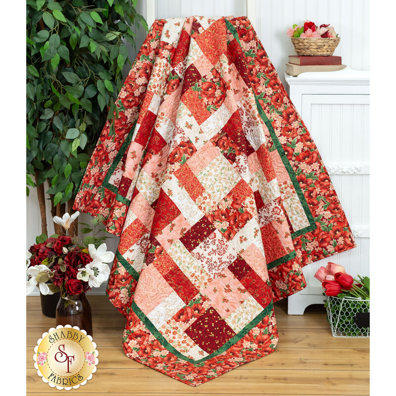 A head on shot of the completed quilt in bright red, pink, crimson, and white, artfully draped over a ladder and staged with coordinating house plants and baskets of flowers.