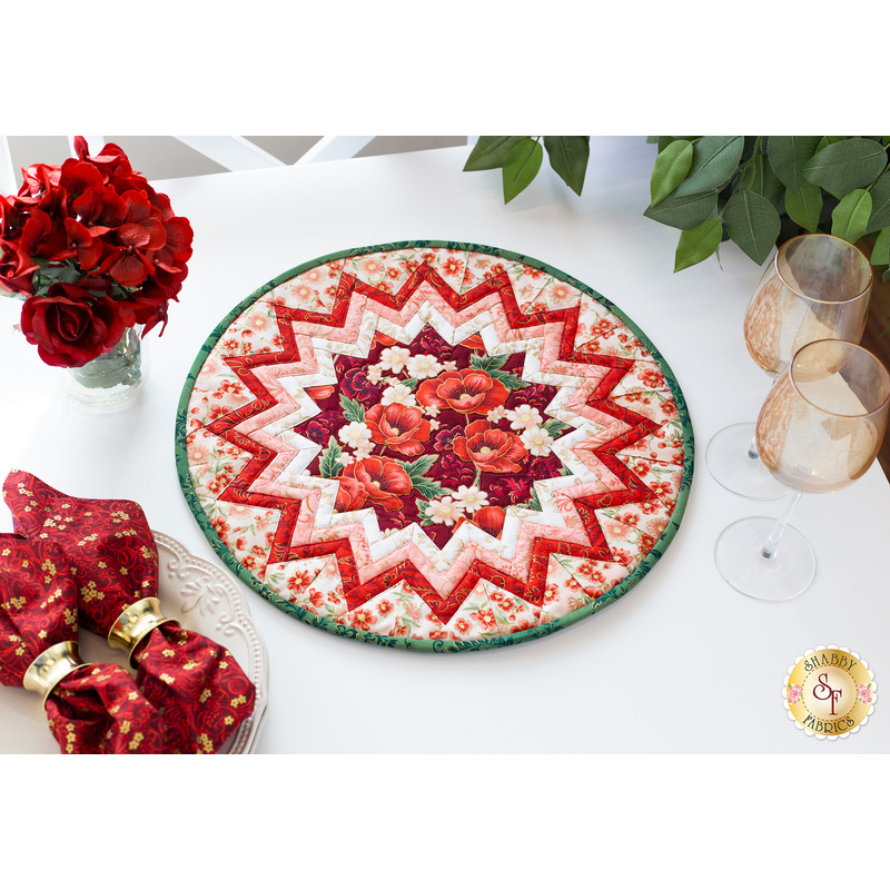 A shot of the completed Point of View Folded Star Table Topper in bright red, pink, white, and green fabrics from the Poppy Hill collection. The Topper is staged on a white table with red roses, red cloth napkins, wine glasses, and a ficus.