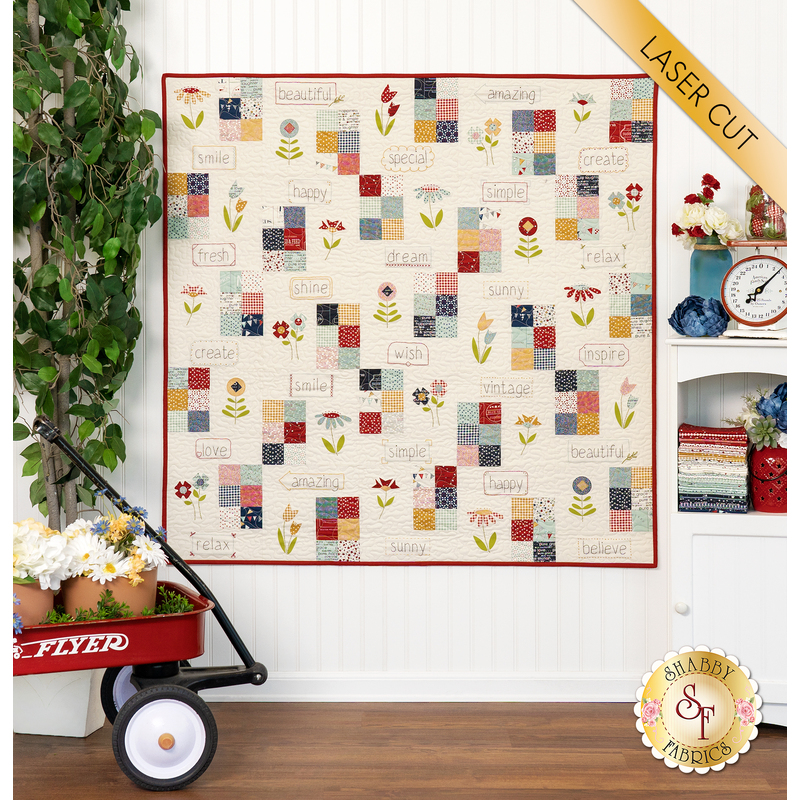 A playful beige quilt hung on a white wall next to a wagon full of flowers