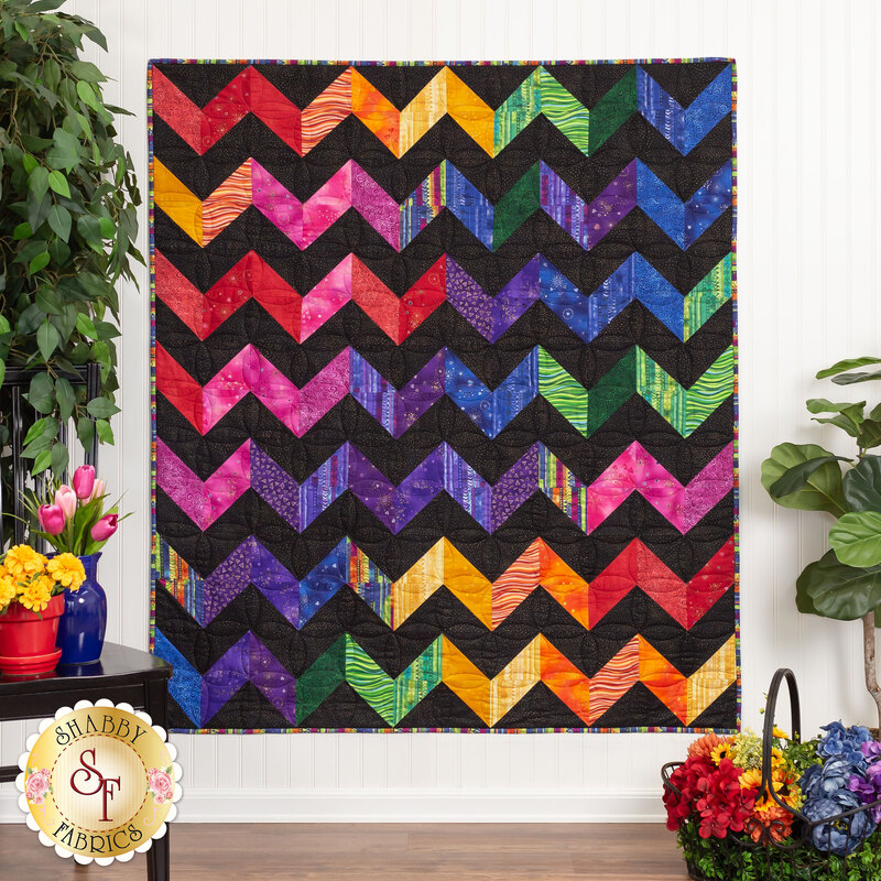 The completed Charming Chevrons Quilt in contrasting black and rainbow fabrics,, hung on a white paneled wall and staged with coordinating flowers and houseplants. A basket of rainbow florals can be seen in the lower right corner.