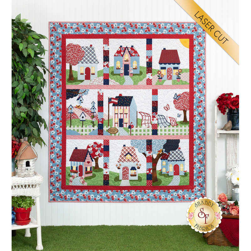The completed Welcome Home In Summer quilt, colored in gentle red, white, and blue. The quilt is hung on a white paneled wall and staged with coordinating greenery and decor. A yellow banner in the top right corner reads 