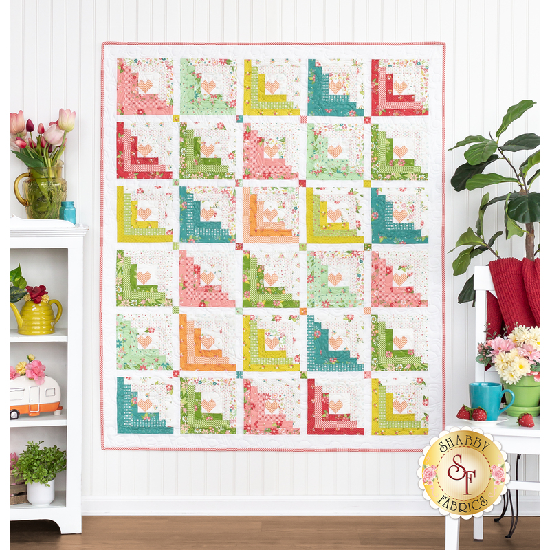 The completed Hearts At Home II Quilt in white and playful colors like lime green, orange, cherry red and pink, and teal blue. Hung on a white paneled wall, the quilt is staged with coordinating furniture and plants, flowers, and ceramic decor.