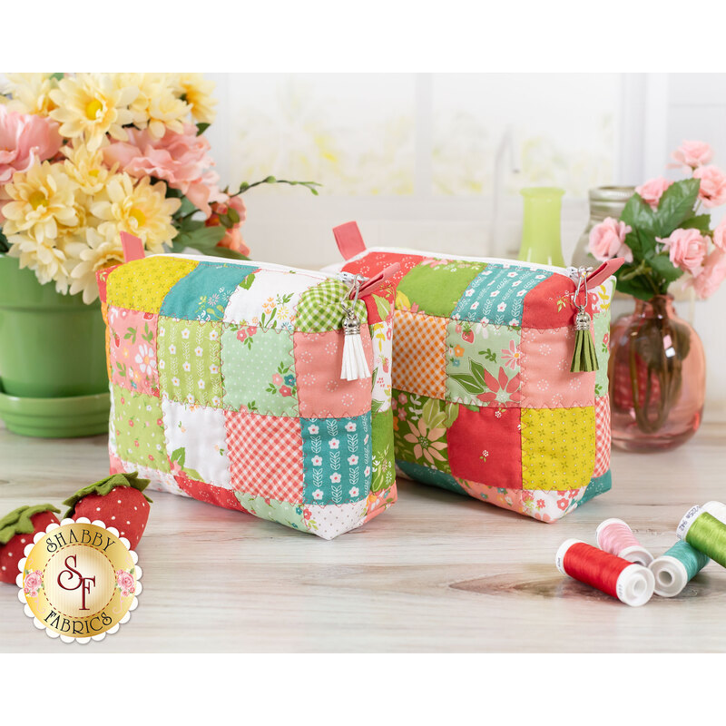 The two completed mini charm bags in playful shades of lime, orange, teal, cherry red, and white. The bags are staged on a light wooden counter with coordinating spools of thread and flowers.