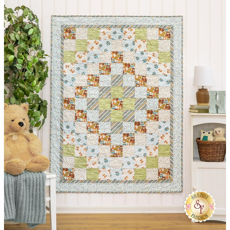The completed quilt in pastel blues, greens, and earthy browns, hung on a white paneled wall. The quilt is staged with coordinating decor and stuffed animals.