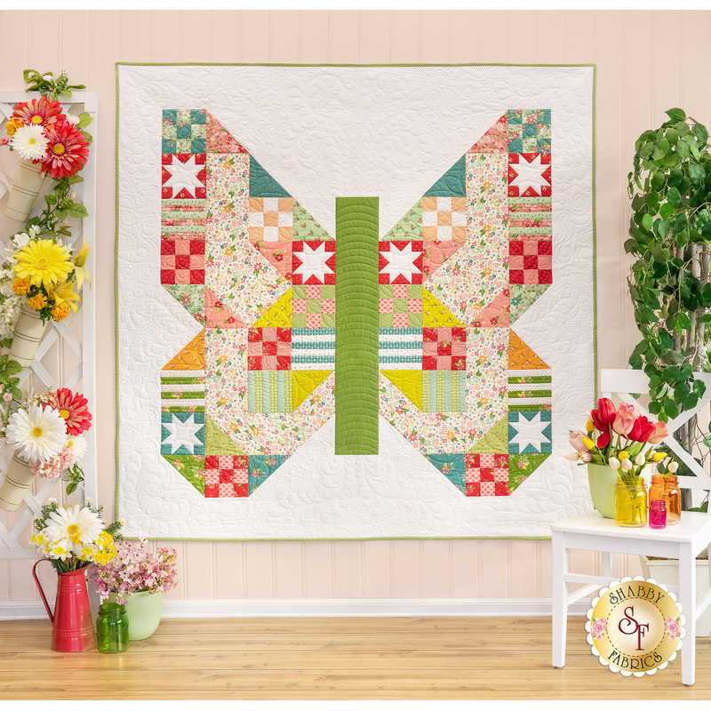 The completed Butterfly Patch Quilt in white and bright colors from Moda's Strawberry Lemonade collection; it is hung on a light blue paneled wall and staged with coordinating decor and flowers.