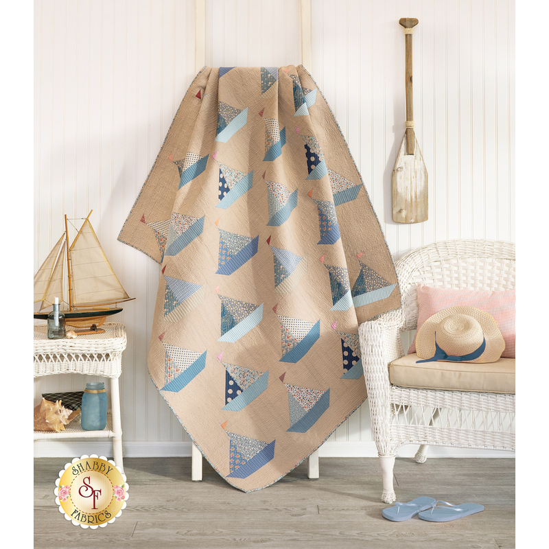 The completed Sailboat quilt in sandy tan and shades of dusty blue, sky blue, and denim. Artfully draped over a white ladder, the quilt is staged with coordinating white furniture and nautical paraphernalia, including a lovely model sailboat.