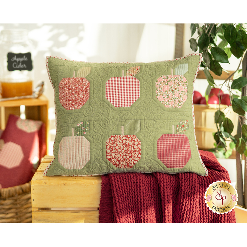 The completed Apple Cider Pillow, staged on a rustic wooden crate, draped with a red knit blanket that complements the pillow. In the background, coordinating decor and furniture are staged with the pillow.