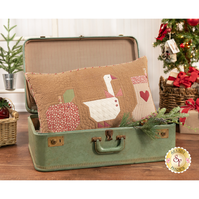 A shot of the completed Winter Pillow in brown. The pillow sits in a vintage green suitcase on a wooden countertop, staged with baskets and Christmas decor like trees and presents.