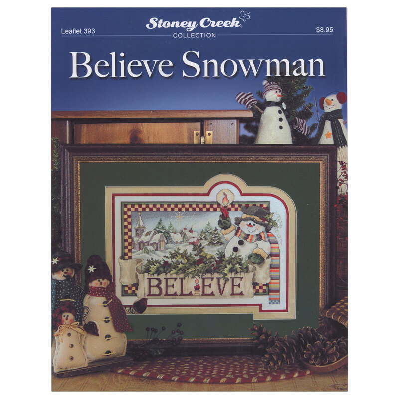 Front of cross stitch pattern showing the finished design framed and displayed on a table with pinecones and snowman figurines