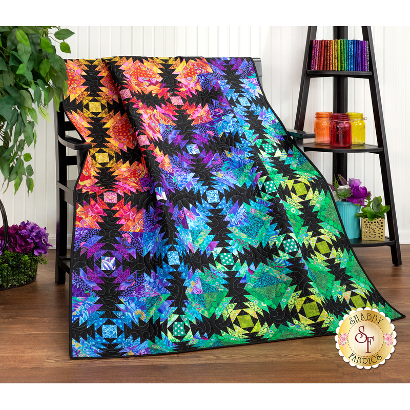 The completed Pineapple quilt in a rainbow of colors and prints, artfully draped over a black bench and staged with coordinating rainbow decor like fabric, jars, and flowers.